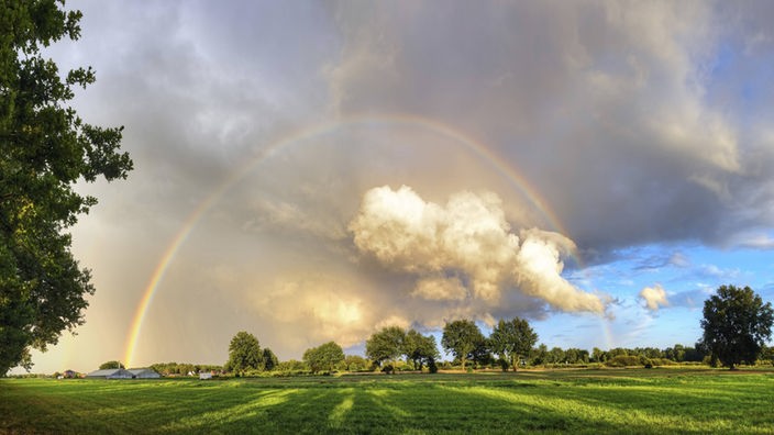 Eine tiefe Wolke mit Regenbogen über einer weiten Grünfläche mit Bäumen am Horizont.