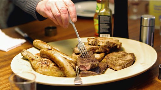 Eine Hand mit Gabel sticht in ein gebratenes Fleischstück, welches auf einem Teller voller verschiedener Fleischstücke und Würste liegt.