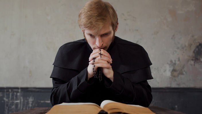 Ein junger Priester sitzt mit gefalteten Händen vor einem aufgeschlagenen Buch.