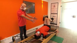 Physiotherapeut mit Patient, der auf einem Gerät liegt für eine Pilates-Rückenübung.