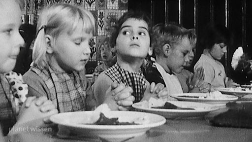 Schwarzweißaufnahme von Kindern, die mit gefalteten Händen am Tisch vor ihren Essenstellern sitzen.