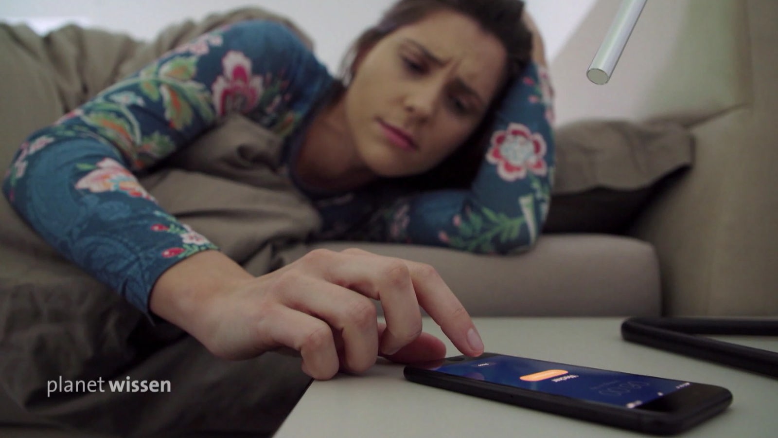 Eine junge Frau liegt im Bett und schaut genervt auf ihr Handy.