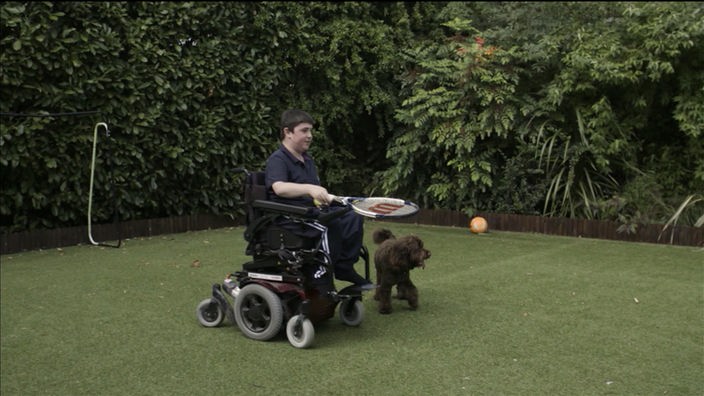 Ein junger Mann in einem Rollstuhl mit Tennisschläger auf einem Rasen im Garten.