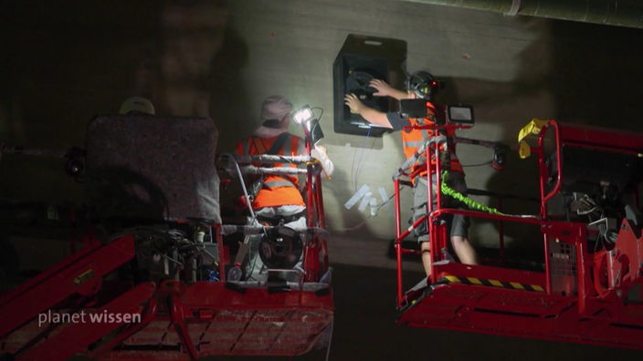 Zwei Personen auf Hubkränen öffnen einen Glasfaserschacht an einer Brücke.