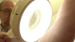 Ein Arzt betrachtet bei einem Hautkrebs-Screening den nackten Arm eines Patienten mit einer Leuchtlupe.
