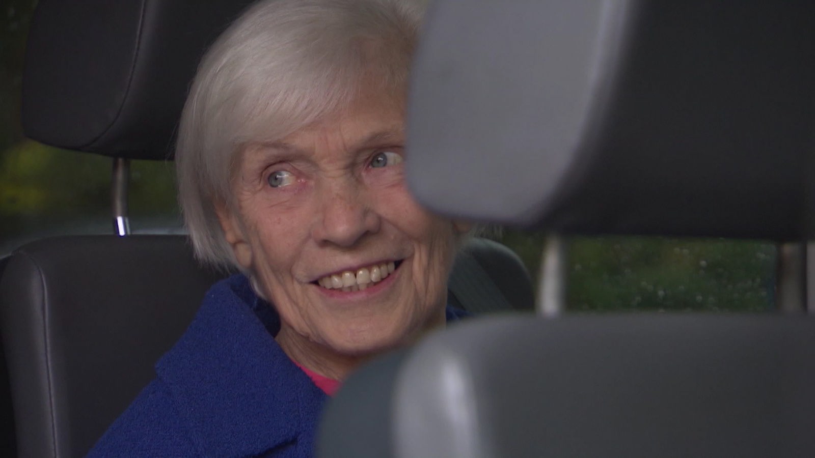 Eine ältere Pflegebedürftige sitzt lächelnd im Auto.