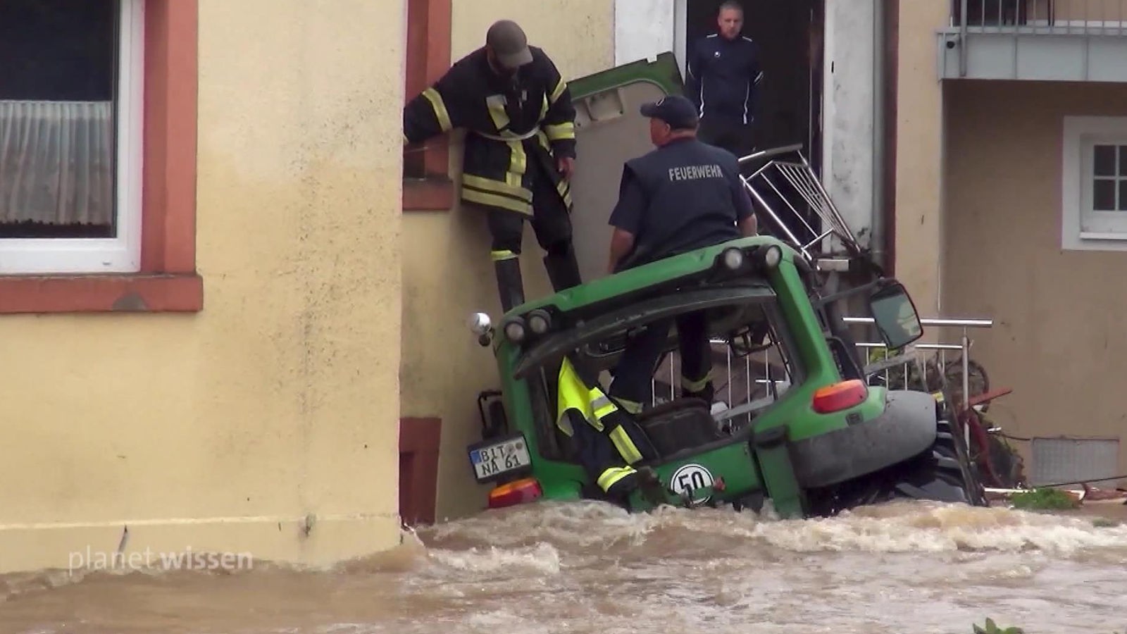 Feuerwehrmänner stehen auf einem Traktor an einer Hauswand, um sie die riesigen Fluten eines Hochwassers.