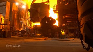 In einer Stahlwerkhalle hängt ein riesiger Schmelzofen an einem Träger, im hellen Licht heißer Flammen.