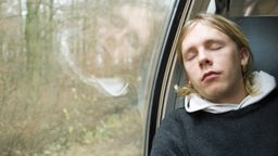 Ein Mann sitzt mit geschlossenen Augen in einem Zug am Fenster.