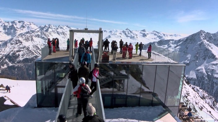 Menschen stehen auf dem Flachdach eines Gebäudes und genießen das Bergpanorama im Sonnenschein.