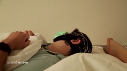 Ein junger Mann mit angelegter EEG-Haube liegt in einem Bett.