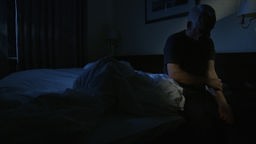 Ein älterer Mann sitzt im Dunkeln auf dem Bettrand und fasst sich an den Arm.