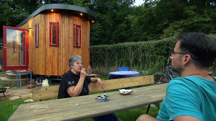 Eine Frau und ein Mann sitzen an einem Gartentisch vor einem Tiny House.
