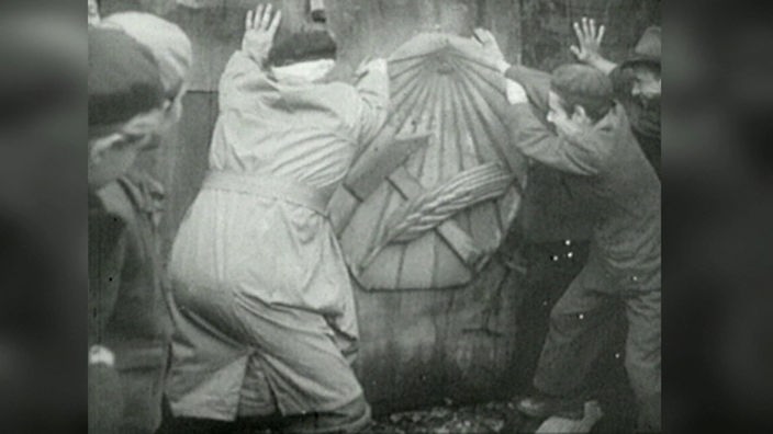 Schwarzweißaufnahme: Männer reißen ein sowjetisches Emblem mit Hammer und Ähre von einer Wand.