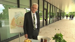 Der baden-württembergische Ministerpräsident Kretschmann steht vor einer Wandtafel mit einer Karte der Dialektregionen von Baden-Württemberg.