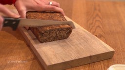Ein dunkles Brot wird von Hand auf einem Holzbrett angeschnitten.