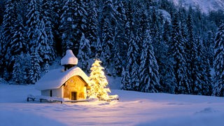 Schneebedeckte Kapelle mit beleuchtetem Tannenbaum daneben