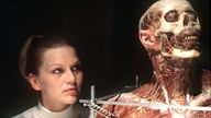Eine blonde Frau neben einer präparierten menschlichen Leiche.