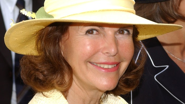 Silvia von Schweden lächelt in die Kamera. Sie trägt ein gelbes Kostüm und einen passenden Hut.