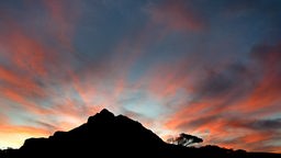 Sonnenuntergang hinter einem Berg im "Table Mountain National Park". Der Berg ist schwarz, der Himmel in verschiedenen Blautönen und die Wolken orangerot gefärbt