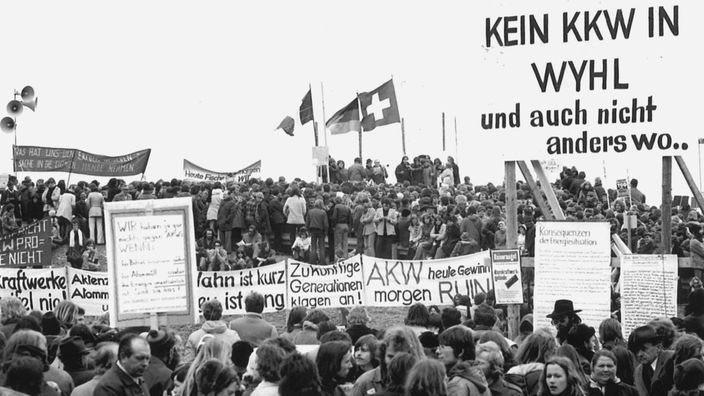 Schwarzweißaufnahme: Demonstration Ostern 1975 in Wyhl gegen das geplante Kernkraftwerk Süd mit Holzschild: 'Kein KKW in Wyhl und auch nicht anderswo'.