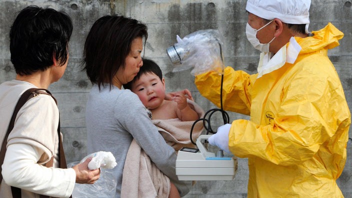 Ein Mann in Schutzkleidung überprüft mit einem Gerät die Strahlung von einer Mutter und ihrem Baby