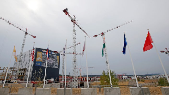 Fusionsreaktor ITER im Bau: Ein großes Gebäude umgeben von Kränen. Im Vordergrund die Flaggen der Partnernationen.