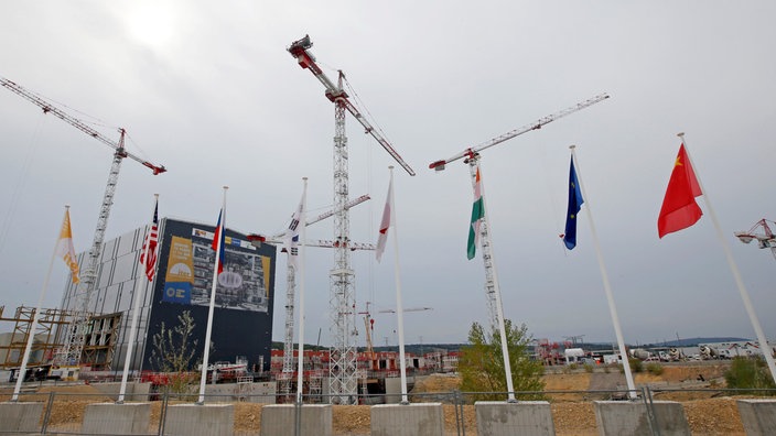 Fusionsreaktor ITER im Bau: Ein großes Gebäude umgeben von Kränen. Im Vordergrund die Flaggen der Partnernationen. | Bildquelle: REUTERS/Jean-Paul Pelissier Fusionsreaktor ITER im Bau: Ein großes Gebäude umgeben von Kränen. Im Vordergrund die Flaggen der Partnernationen.