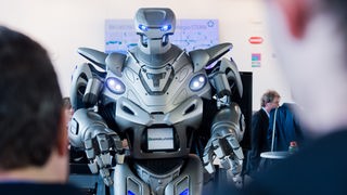 Ein futuristisch aussehender Roboter auf der Hannover Messe über die Schulter zweier Besucher fotografiert. 