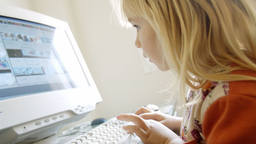 Ein kleines Mädchen schaut gespannt auf einen Computerbildschirm und hält die Händen über einer Tastatur.