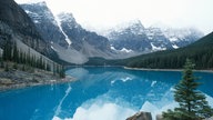 Ein waldgesäumter blauer See. Im Hintergrund schneebedeckte Bergkuppen