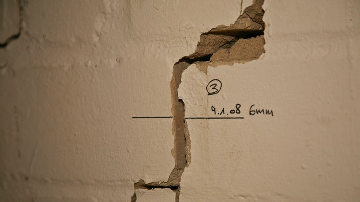 Riss in einer weiß getünchten Wand. Rechts neben dem Riss ist mit Filzstift die Aufschrift '6 mm' geschrieben.