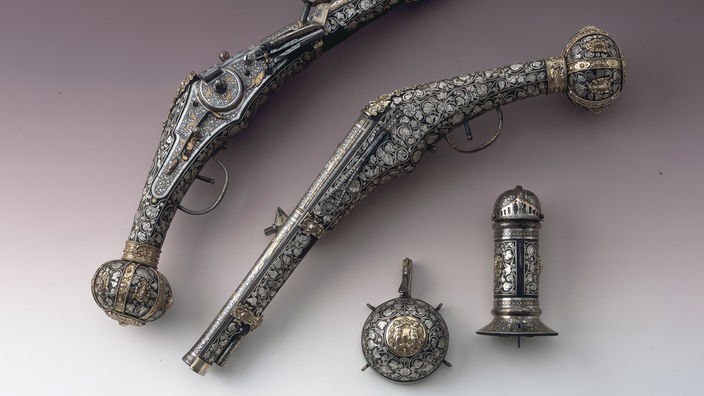 Zwei reich verzierte Pistolen und ein Fläschchen liegen vor einfarbigem Hintergrund