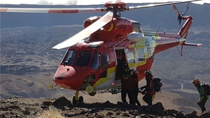 Ein Rettungshubschrauber steht am Rand eines Vulkans. Von rechts kommen Menschen, die in den Heli einsteigen wollen.