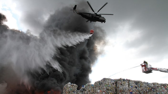 Ein Hubschrauber lässt aus einem Behälter Wasser über einem brennenden Müllberg ab.