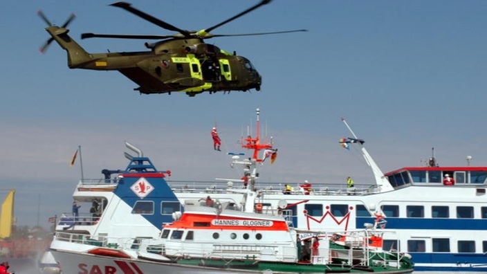 Ein Hubschrauber schwebt über zwei Schiffen.  Eine Person seilt sich aus dem Hubschrauber ab