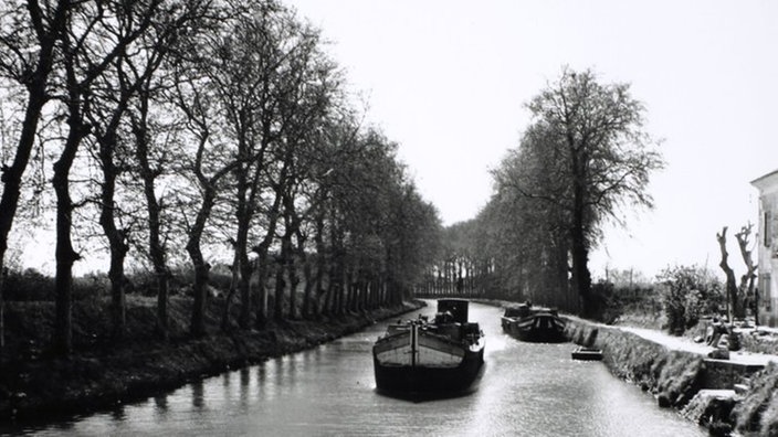 Das Schwarzweiß-Foto zeigt einen gradlinigen Kanal, dessen Ufer Wege mit Bäumen gesäumt sind. Ein Schiff befährt den Kanal. Eines liegt am Ufer. | Bildquelle: akg Das Schwarzweiß-Foto zeigt einen gradlinigen Kanal, dessen Ufer Wege mit Bäumen gesäumt sind. Ein Schiff befährt den Kanal. Eines liegt am Ufer.