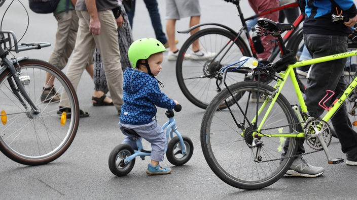Kleines Kind mit Laufrad und Helm inmitten von Fußgängern und Fahrradfahrern.