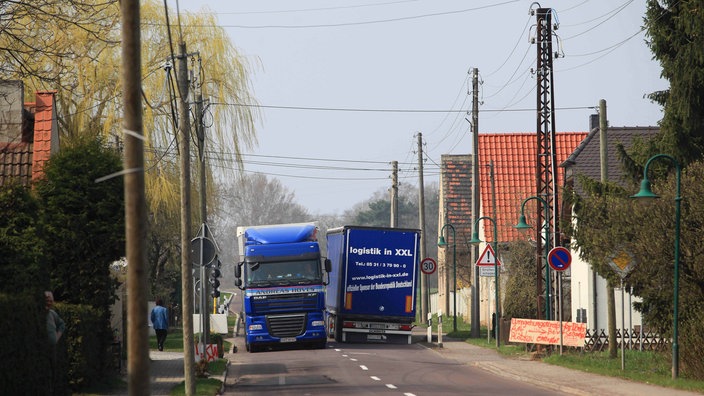 Zwei Lkws zwängen sich über eine enge Dorfstraße aneinander vorbei.