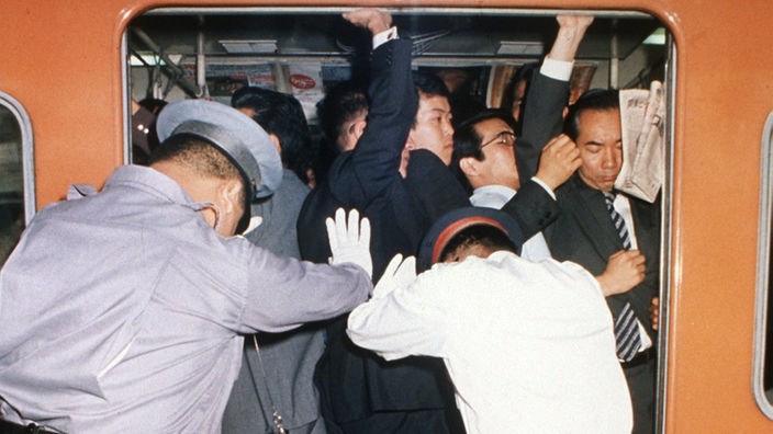 Die geöffnete Tür eines vollbesetzten U-Bahn-Waggons. Von außen drücken Männer in Uniformen und weißen Handschuhen die Fahrgäste in den Wagen