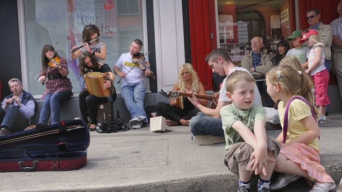 Neben einem Café spielen Straßenmusiker. Kinder sitzen auf dem Trottoir. Erwachsene sitzen gemütlich vor dem Café und hören der Musik zu. 