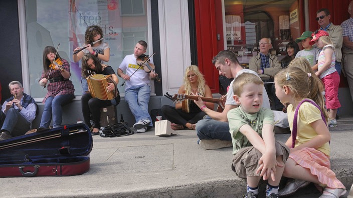 Neben einem Café spielen Straßenmusiker. Kinder sitzen auf dem Trottoir. Erwachsene sitzen gemütlich vor dem Café und hören der Musik zu. | Bildquelle: Imago Neben einem Café spielen Straßenmusiker. Kinder sitzen auf dem Trottoir. Erwachsene sitzen gemütlich vor dem Café und hören der Musik zu.