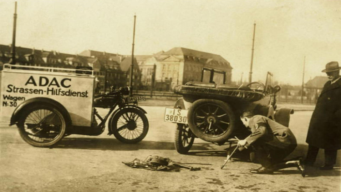 Schwarzweiß-Foto: Ein ADAC-Mitarbeiter hilft 1925 einem Autofahrer mit Panne. Links im Bild ein Motorrad mit Anhänger, auf dem die Aufschrift 'ADAC-Straßenhilfsdienst' zu sehen ist.