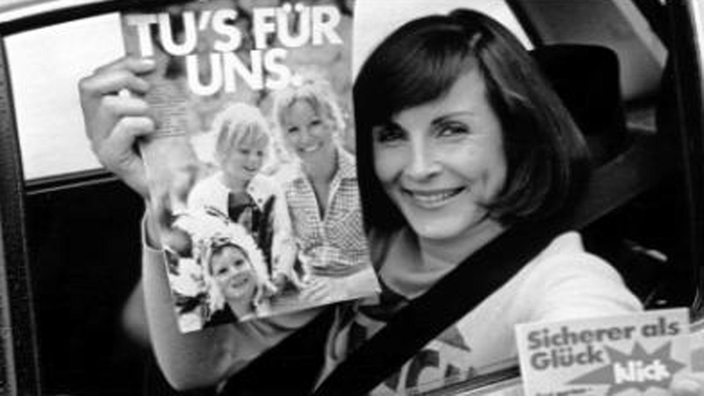 Schwarzweiß-Foto: Eine Frau sitzt in einem Auto, schaut aus dem Fenster und hält ein Plakat mit der Aufschrift 'Tu's für uns' in die Kamera. Auf der Autotür steht 'Erst gurten, dann starten'.