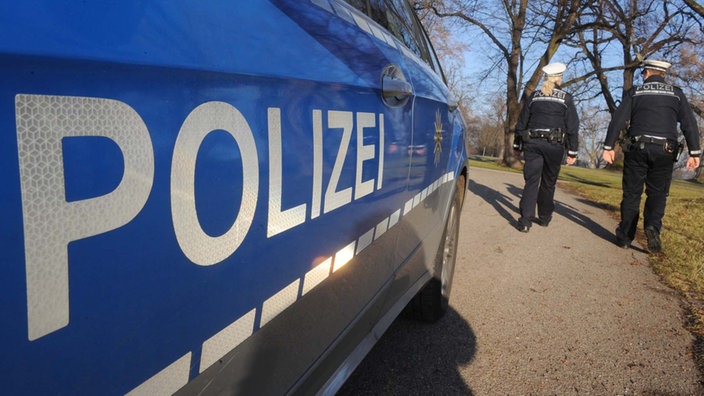 Zwei Polizisten laufen einen Weg entlang, im Vordergrund steht das Polizeiauto. | Bildquelle: dpa/Franziska Kraufmann Zwei Polizisten laufen einen Weg entlang, im Vordergrund steht das Polizeiauto.