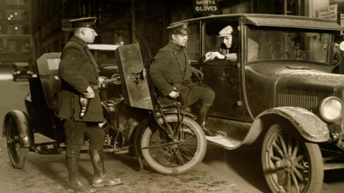 Schwarzweiß-Foto: Zwei Polizisten haben gerade mit einem Motorrad mit Beiwagen einen Autofahrer angehalten. Der Autofahrer reicht den Polizisten aus dem geöffneten Fenster seine Papiere.