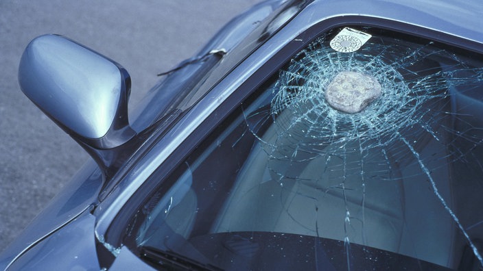 Ein etwa faustgroßer Stein in der Frontscheibe eiens Autos. Die Scheibe ist lediglich angerissen. | Bildquelle: Mauritius Ein etwa faustgroßer Stein in der Frontscheibe eiens Autos. Die Scheibe ist lediglich angerissen.