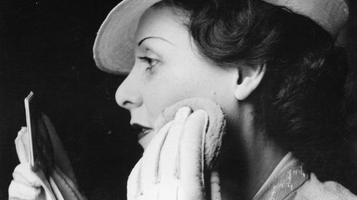 Schwarzweiß-Bild: Eine Frau trägt weiße Lederhandschuhe. Sie hat einen Spiegel in der Hand und schminkt sich.