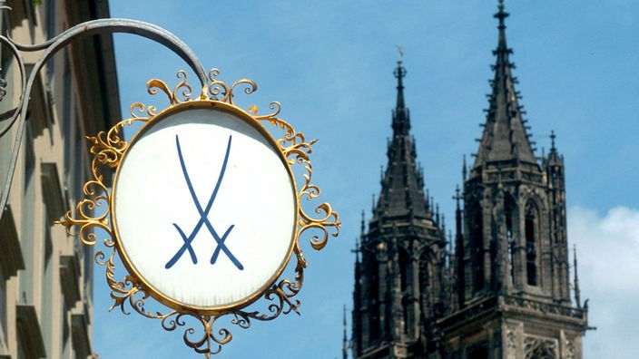 Das Wappen der Porzellanmanufaktur aus Meißen hängt kunstvoll verziert an einem Haus. Im Hintergrund zwei Kirchturmspitzen.