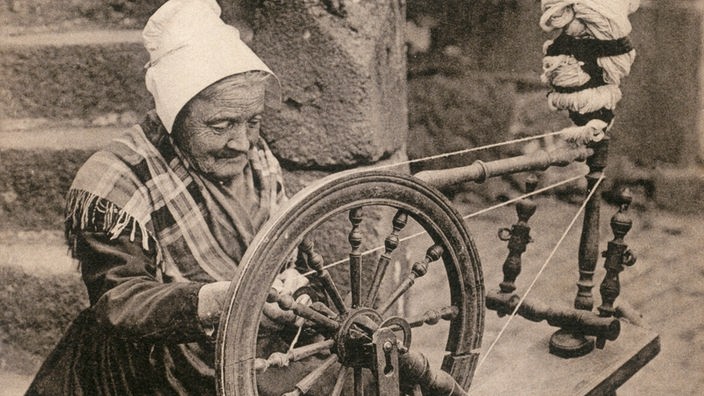 Schwarzweiß-Foto einer alten Frau hinter einem Spinnrad.
